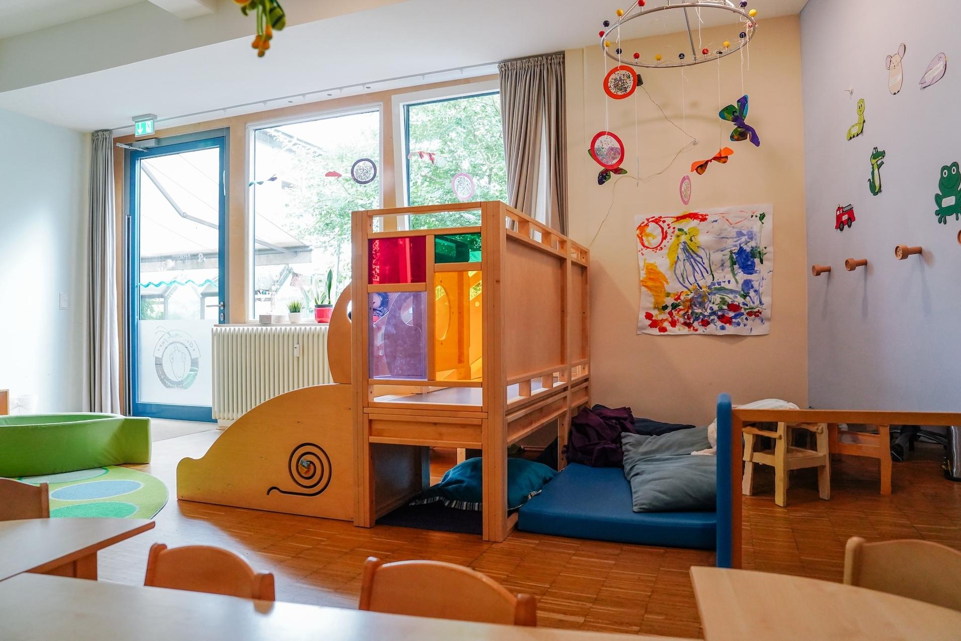 Spielbereich in einem Zimmer mit Holzkonstruktion, bunten Fenstern, Spielmatte und Bildern an der Wand.