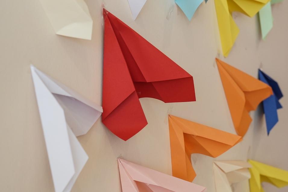 Bunte Papierflieger in unterschiedlichen Farben hängen an einer Wand, mit verschiedenen Designs und Anordnungen.